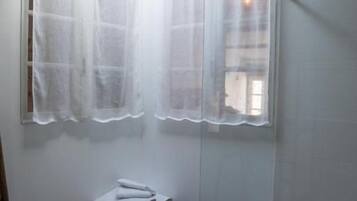 A small bathroom with a shower, a bench, and a window with sheer curtains.