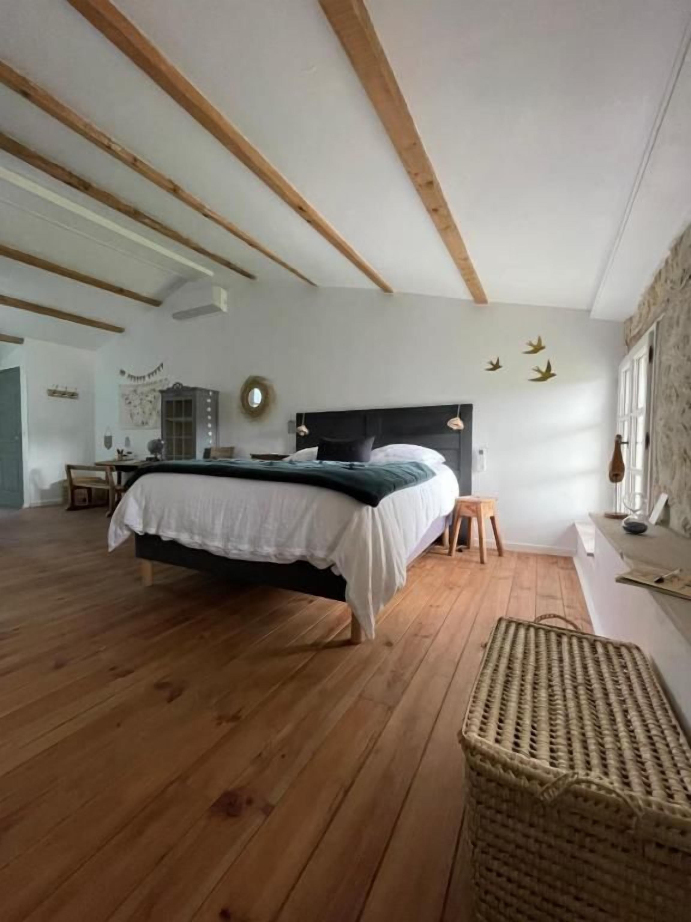 A bedroom with a wooden floor, a bed with a white bedspread, a small stool, and a woven basket.
