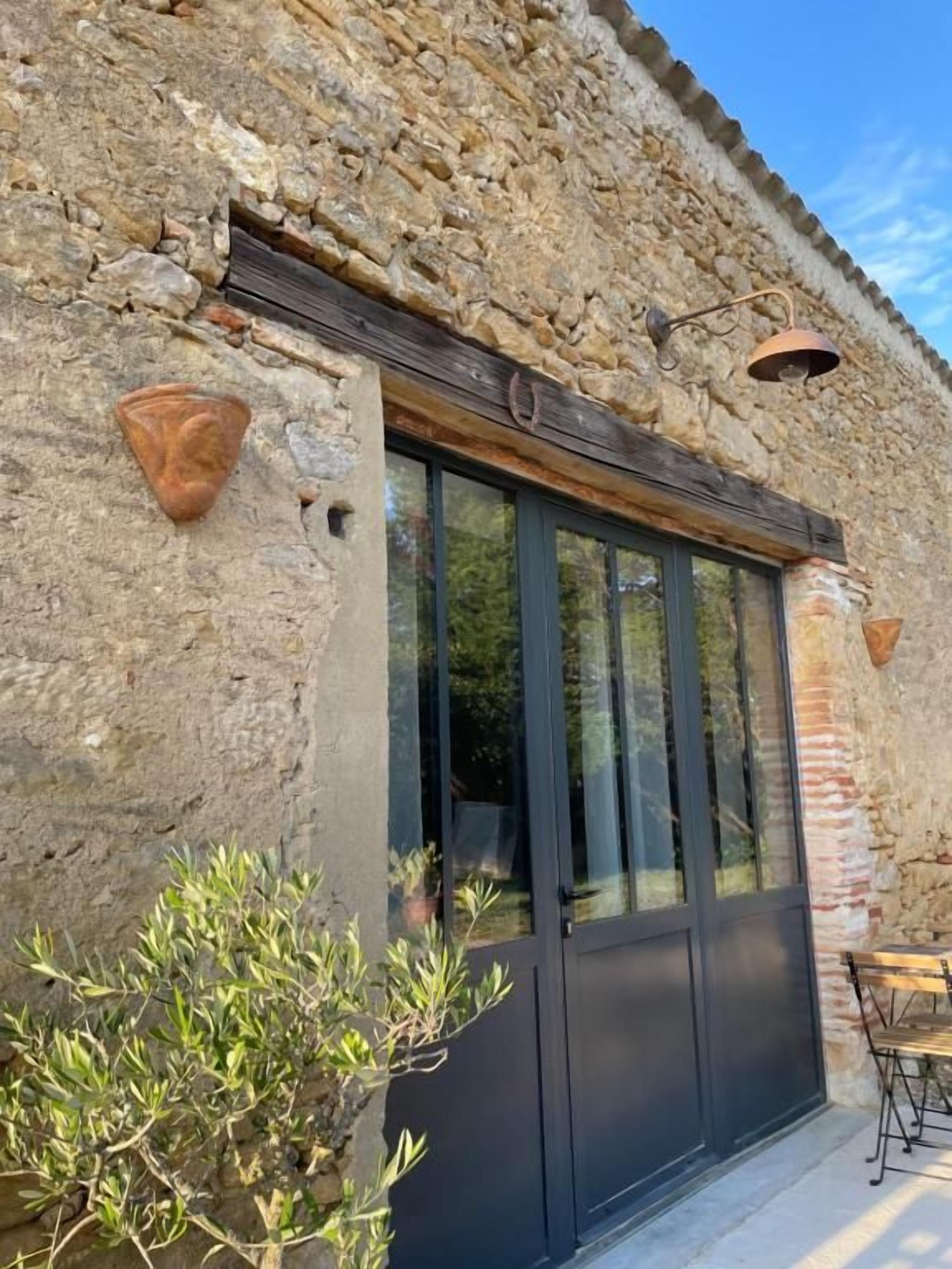 Un bâtiment en pierre doté d’une grande fenêtre et d’une porte, d’une petite table et d’une plante en pot.