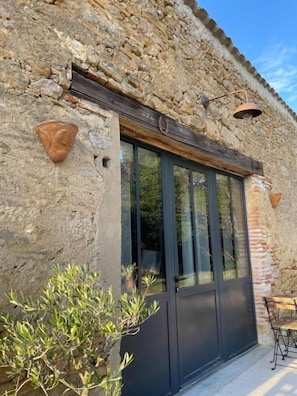 Un edificio de piedra con un ventanal grande y una puerta, una mesita pequeña y una planta en maceta.