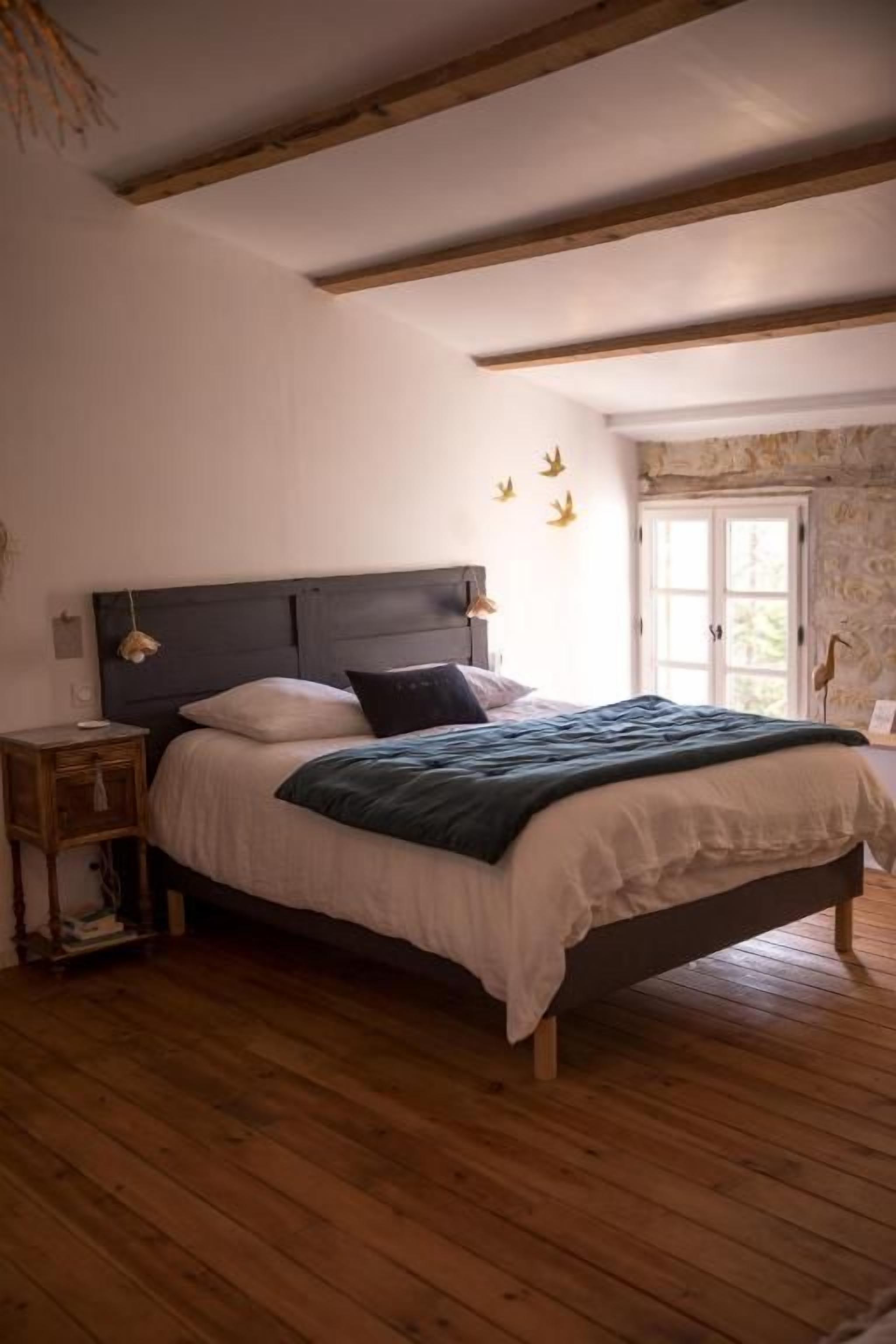 A bedroom with a wooden floor, a dark headboard, a bed with white linens, and a nightstand with a lamp.