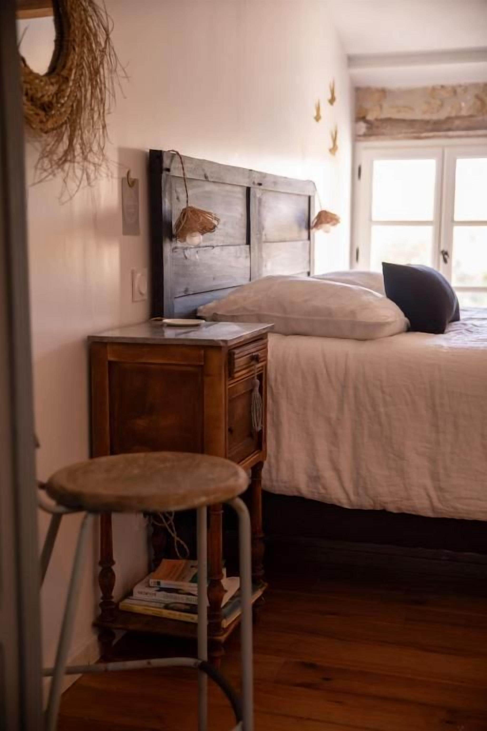 A bedroom with a wooden headboard, a bed with white bedding, a wooden nightstand with books, and a window with curtains.