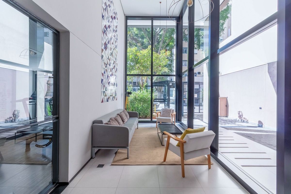 A modern living room with a sofa, chairs, and a dining table. Large windows provide a view of the outdoors.
