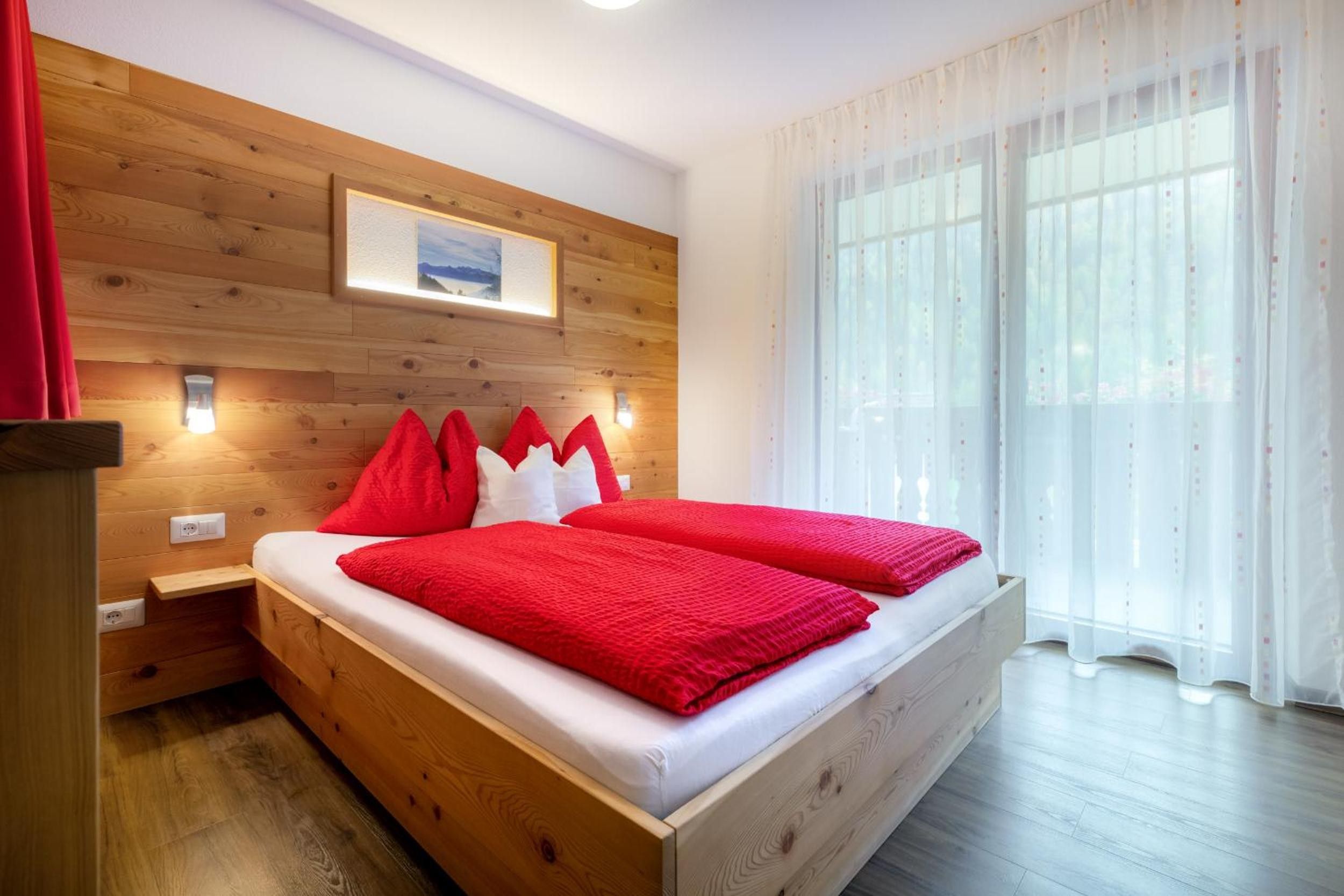 Ein Schlafzimmer mit einem hölzernen Kopfteil, einem Bett mit roter Bettwäsche, einem Nachttisch und einem Fenster mit durchsichtigen Vorhängen.