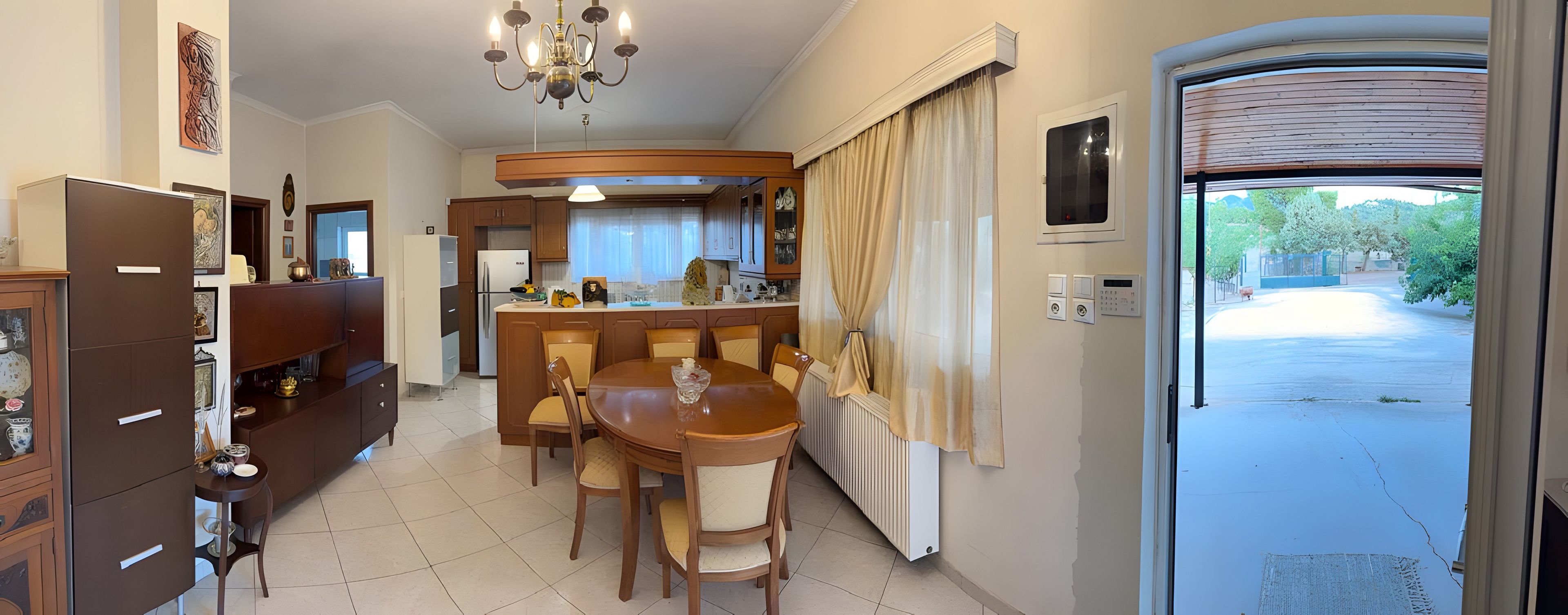 A dining area with a wooden table and chairs, a kitchen in the background, and a view of the outside.