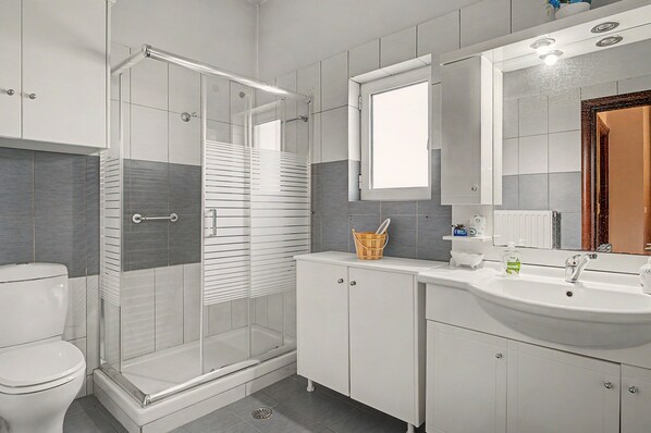 A modern bathroom with a glass shower enclosure, a white toilet, a sink with a mirror above, and a cabinet with a basket.