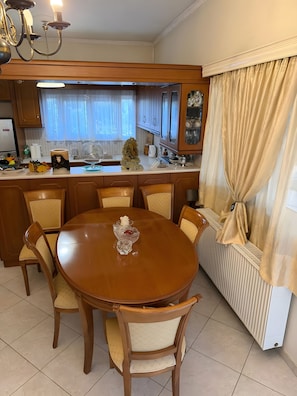 A dining area with a wooden table and chairs, a chandelier, and a window with curtains. - Country House 'Ochra' with Mountain View, Wi-Fi and Air Conditioning (δημοτική ενότητα Λουτρακίου-Περαχώρας)