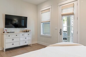 A bedroom with a large flat-screen TV mounted on the wall, a white dresser with drawers, a bed with a white comforter, and two windows with white blinds. - Out of Office Beaches | Lagoon Pool, 7 Bikes, Screened Balcony, Sleeps 14 (Seacrest Beach)