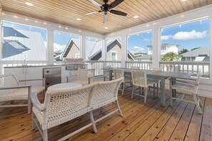 A covered outdoor patio with a grill, wicker furniture, and a view of neighboring houses. - Out of Office Beaches | Lagoon Pool, 7 Bikes, Screened Balcony, Sleeps 14 (Seacrest Beach)