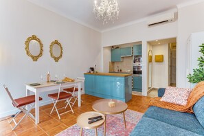 A modern living room with a sofa, a dining table with chairs, a kitchen area with blue cabinets, and a chandelier. - THE LIBERTY AP4510 - Clim - 5-6 min de la plage by (Nice)