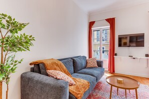 A living room with a grey sofa, a small round table, a red curtain, a television on the wall, and a potted plant. - THE LIBERTY AP4510 - Clim - 5-6 min de la plage by (Nice)