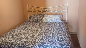 Una cama individual con estructura metálica y colcha estampada floral en una habitación con paredes color melocotón y suelo de madera.