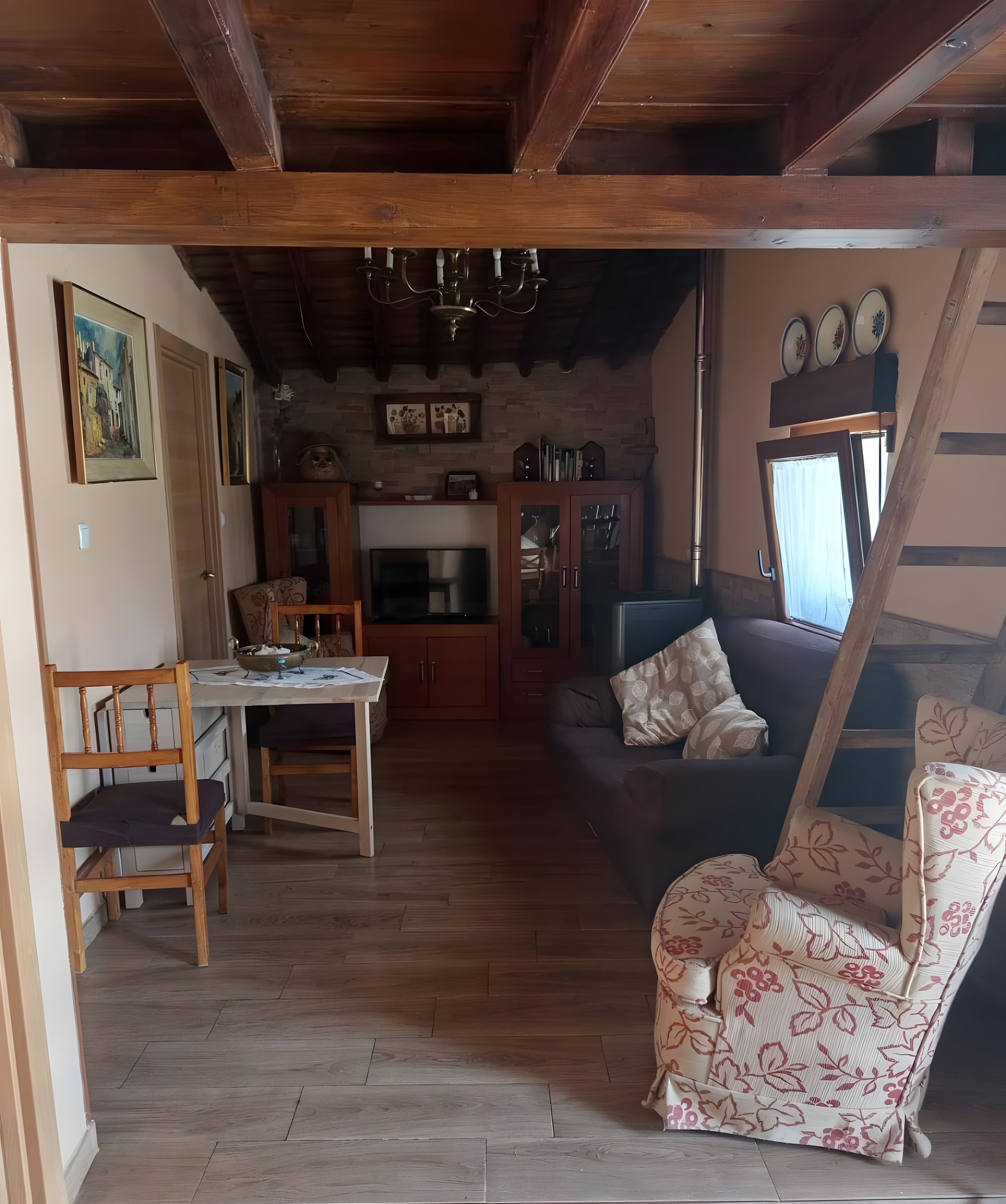 Una sala de estar acogedora con piso de madera, un sofá, un sillón estampado, una mesa de comedor con sillas y una cocina al fondo.