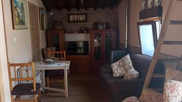 Una sala de estar acogedora con piso de madera, un sofá, un sillón estampado, una mesa de comedor con sillas y una cocina al fondo.