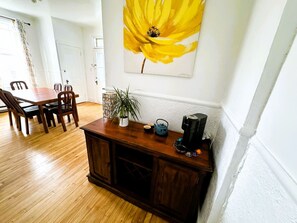 Ein Essbereich mit einem Holztisch und Stühlen, einer Kommode mit einer Pflanze, einer Kaffeemaschine und einem Gemälde einer gelben Blume an der Wand.