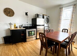 Cocina compacta con una mesa y sillas de madera, horno empotrado, lavadora y un reloj en la pared.