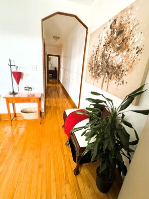 A room with a wooden floor, a bed with a red blanket, a potted plant, a desk with a lamp, and a wall with a textured painting.