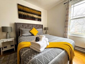 A bedroom with a bed, a bedside table, a lamp, a window with curtains, and a painting on the wall.