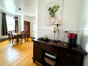 A dining area with wooden furniture, a table lamp, a coffee machine, and a cactus painting on the wall.
