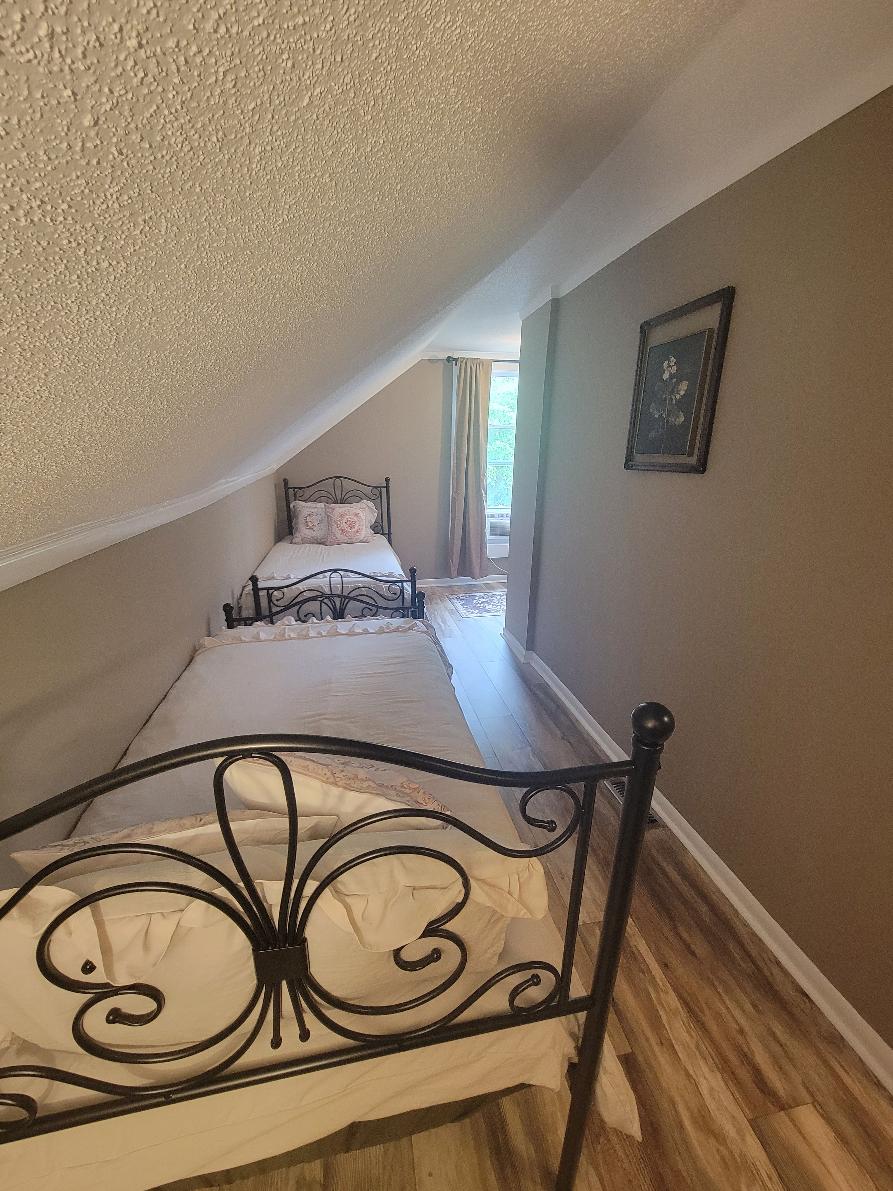 Un dormitorio con techo inclinado, una cama con cabecera decorativa, un cuadro enmarcado en la pared y una ventana con cortinas.