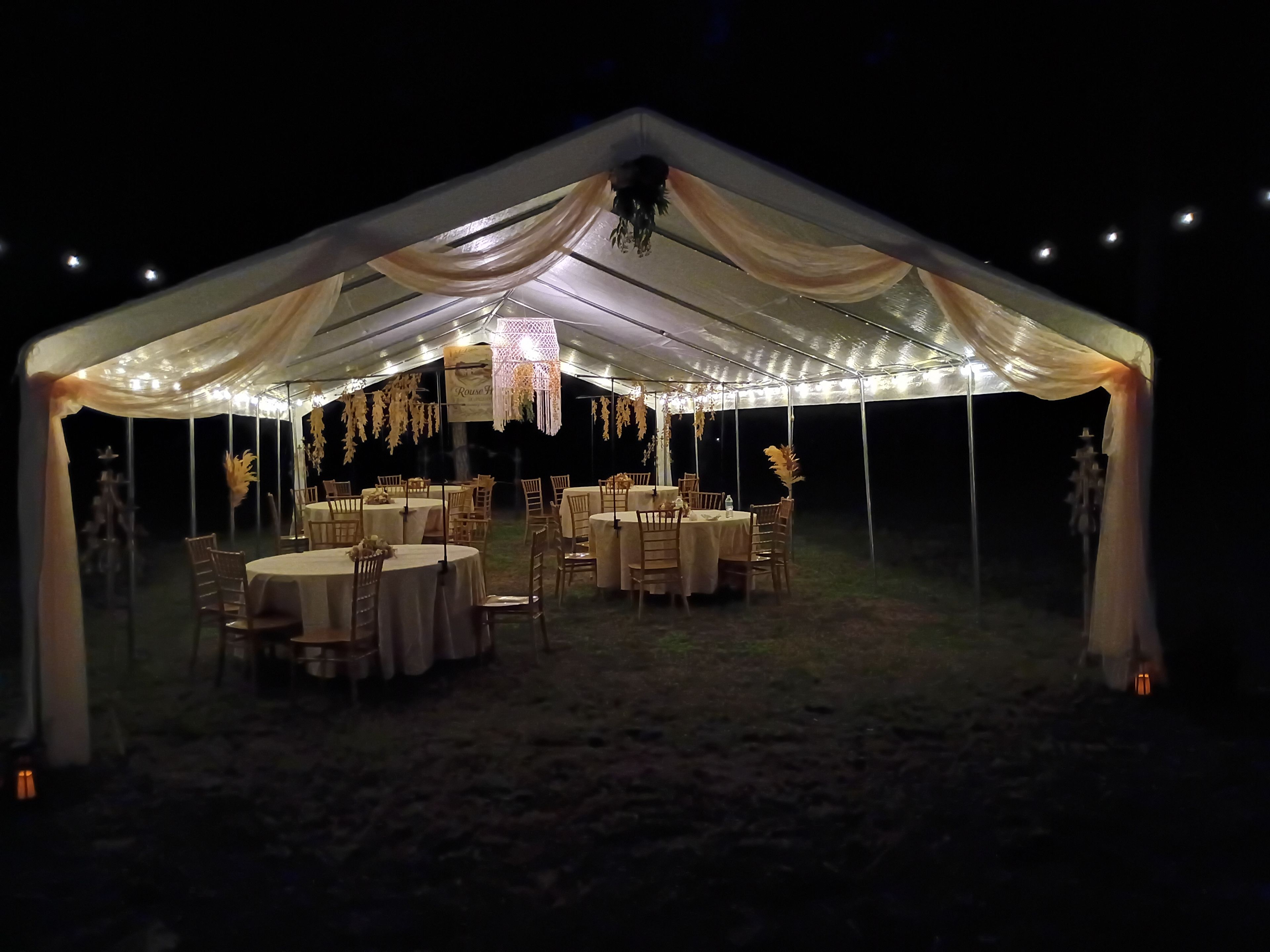 Un espacio al aire libre con carpas, mesas redondas y sillas, iluminado con luces colgantes.