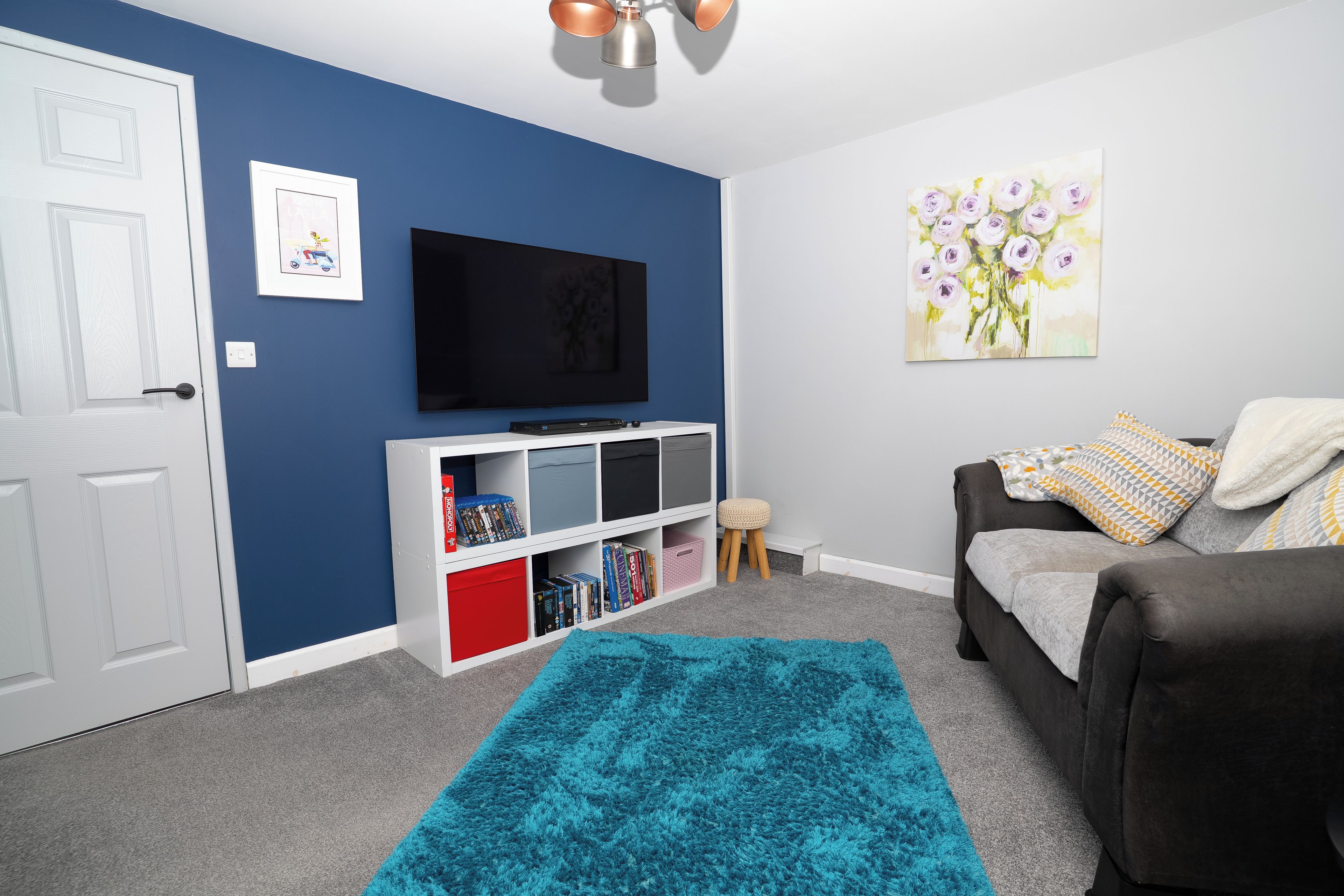 A living room with a blue wall, a flat-screen TV, a bookshelf, a grey sofa, a blue rug, and a painting of flowers.