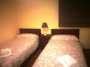 Et hotelværelse med to senge, et natbord med lampe og et vindue med mørklægning.