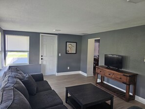 A living room with a sectional sofa, a coffee table, a TV on a wooden stand, and a door leading to another room. - Lovely 2-bedroom house with AC in charming Englewood with Boat slip to Lemon Bay (Englewood)