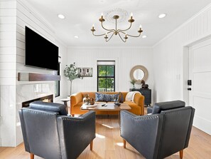 A modern living room with a fireplace, leather chairs, a sofa, and a chandelier. - Historic Swansboro Retreat | Water Views + Backyard with Pizza Oven (Swansboro)