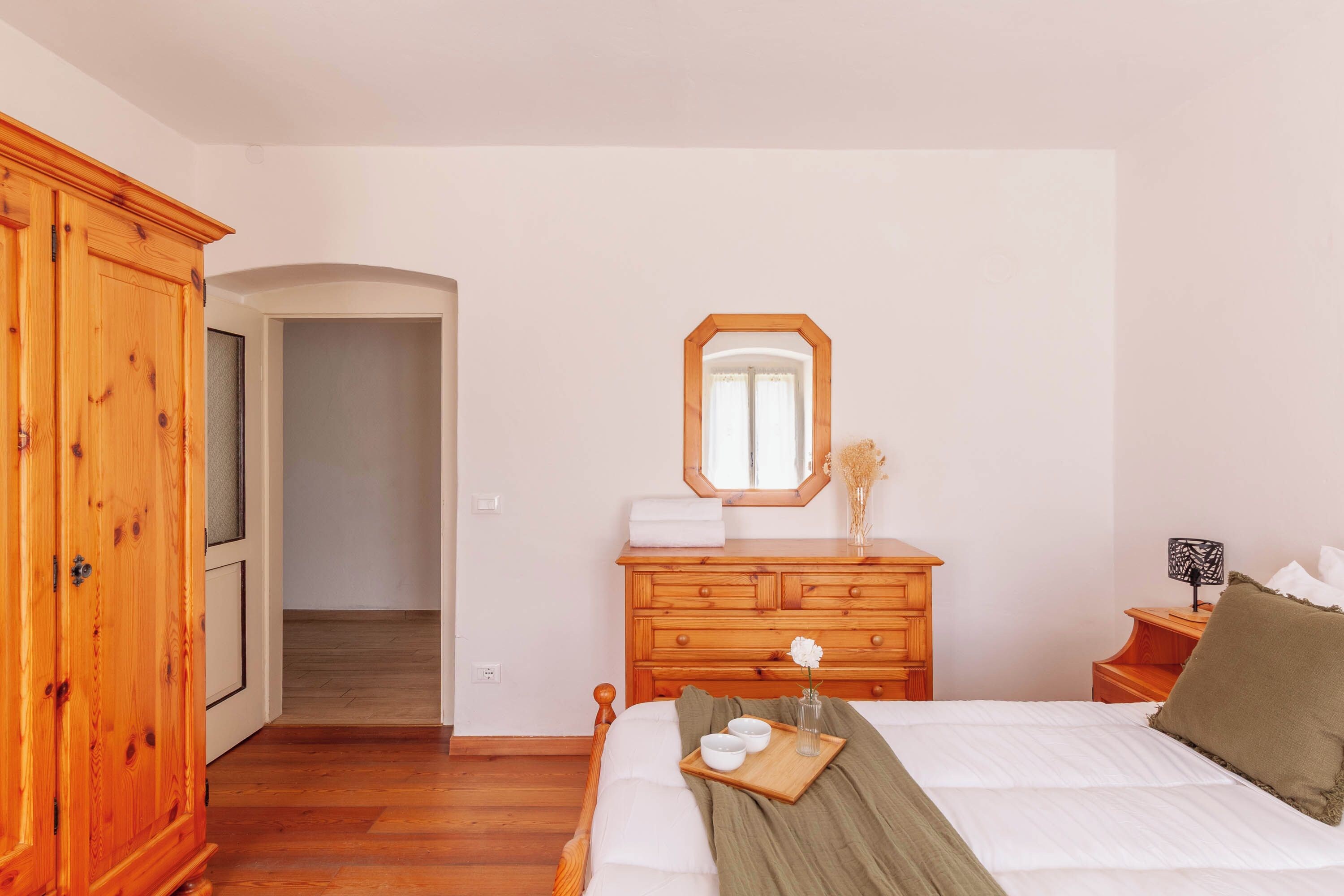 A bedroom with a wooden wardrobe, a bed with a green throw, a dresser with a mirror, and a nightstand with a lamp.
