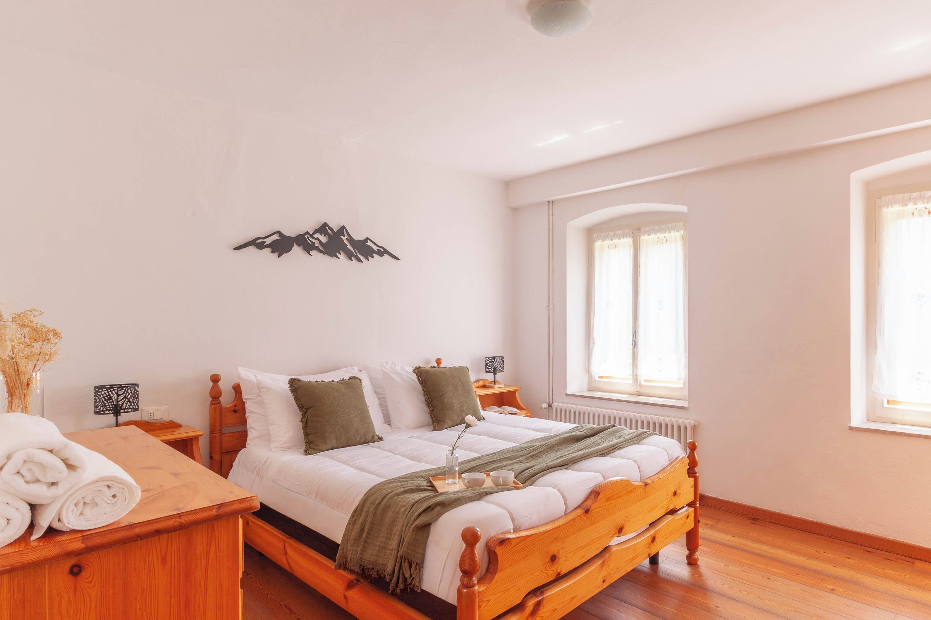 Une chambre avec un lit en bois, du linge de lit blanc, une décoration représentant une montagne au mur, une fenêtre avec des rideaux et une table de chevet avec une lampe.