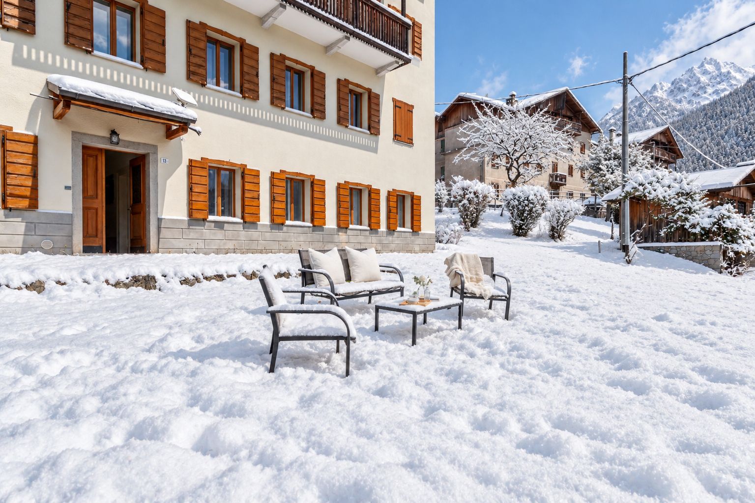 A snow-covered outdoor seating area with chairs and tables, a building with wooden shutters, and snow-covered trees.