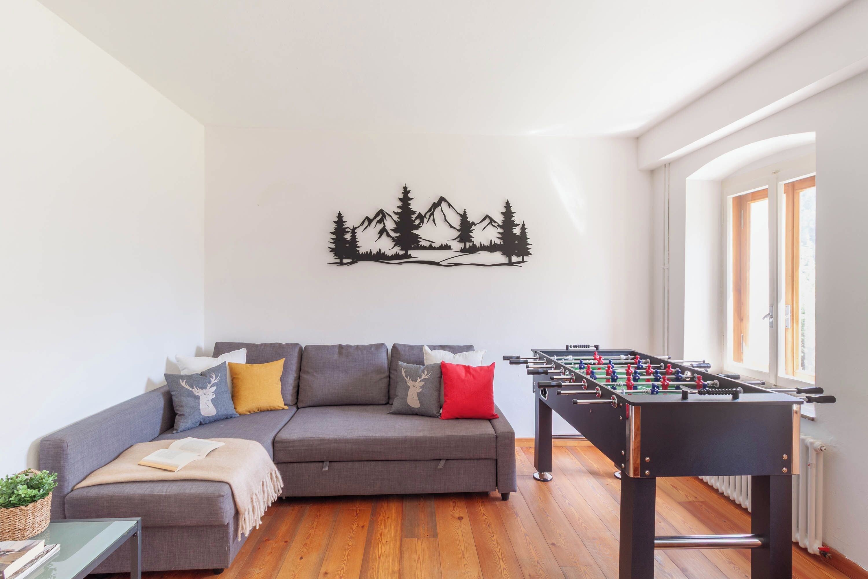 A living room with a grey sofa, a foosball table, and a wall art of mountains.