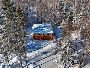 Une cabane dont le toit est recouvert de neige, entourée d’arbres et sous un ciel dégagé.