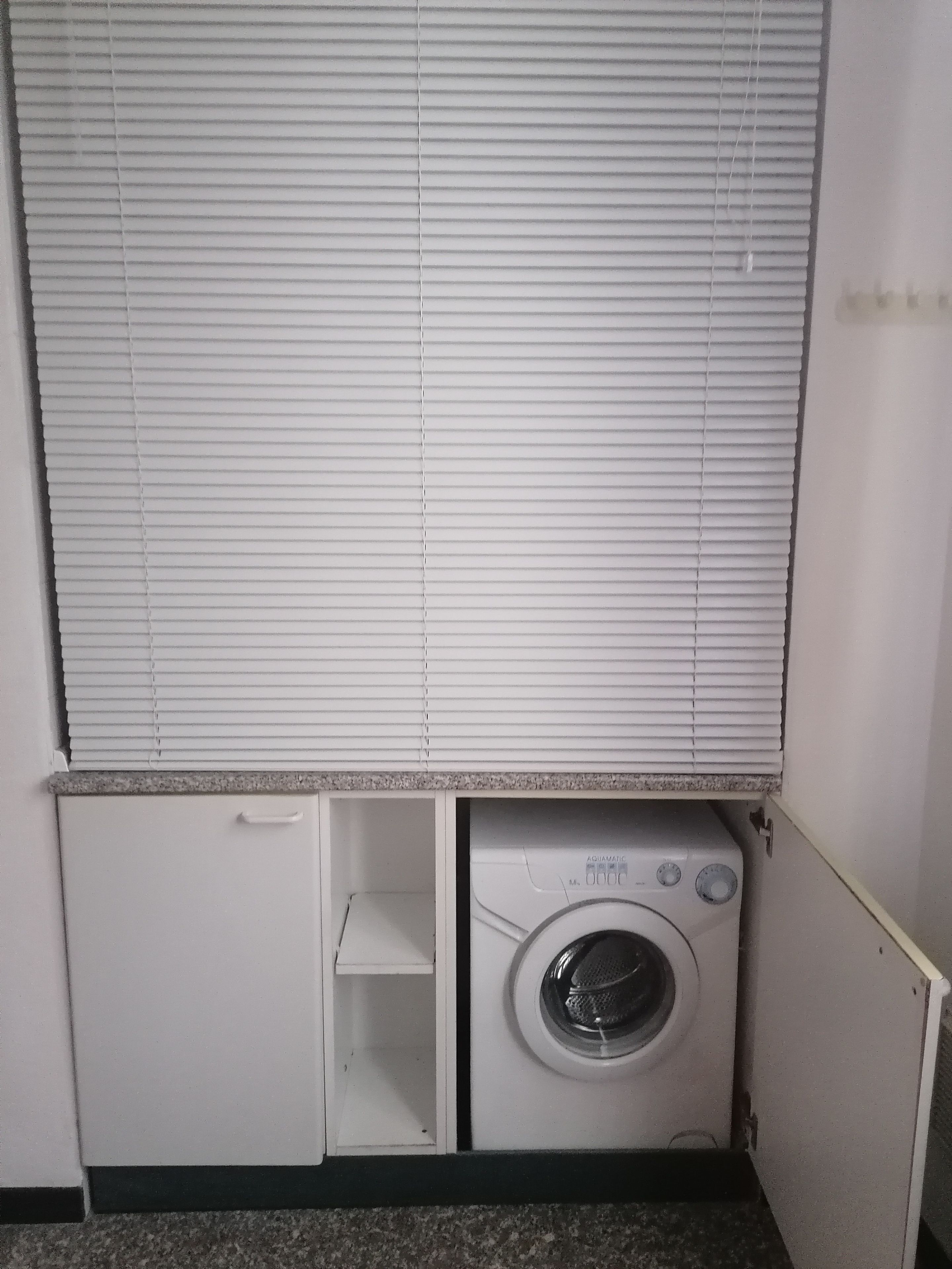 Ein kompakter Hauswirtschaftsraum mit einer Waschmaschine mit Frontlader, einem kleinen Schrank und einem Fenster mit Jalousien.