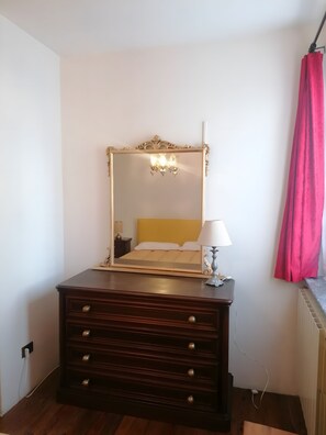 Ein Zimmer mit einem großen Spiegel, einem Bett mit gelber Kopfteil, einer Kommode mit Schubladen, einer Lampe und einem roten Vorhang.