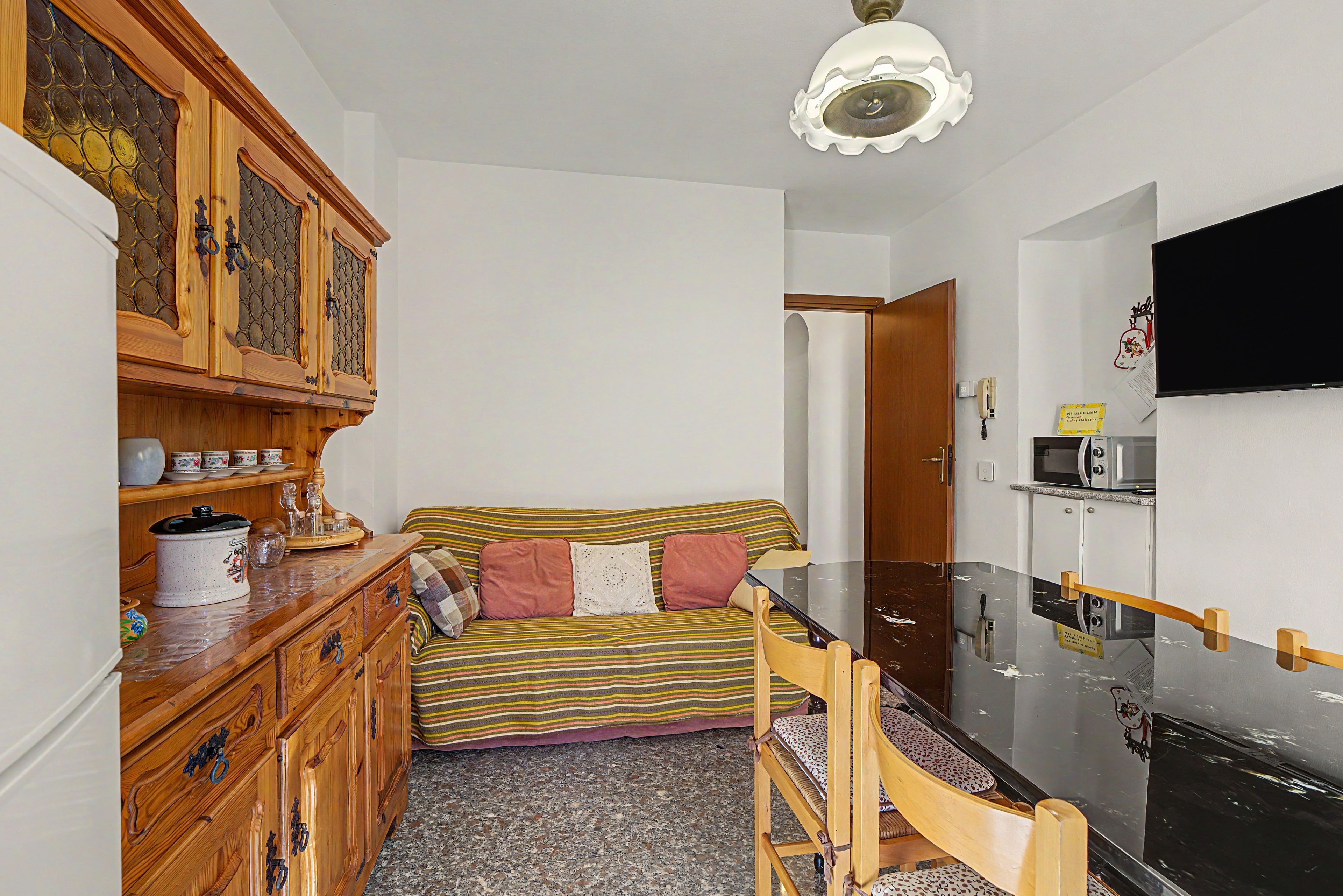 A compact living space with a kitchenette, a dining area with a wooden table and chairs, a sofa with striped cushions, and a television mounted on the wall.