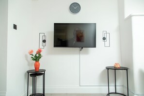 A room with a flat-screen TV mounted on the wall, a vase with flowers on a small table, and another table with a decorative item. - Central Wembley - Apartment - Sleeps 5 - Parking (Wembley)