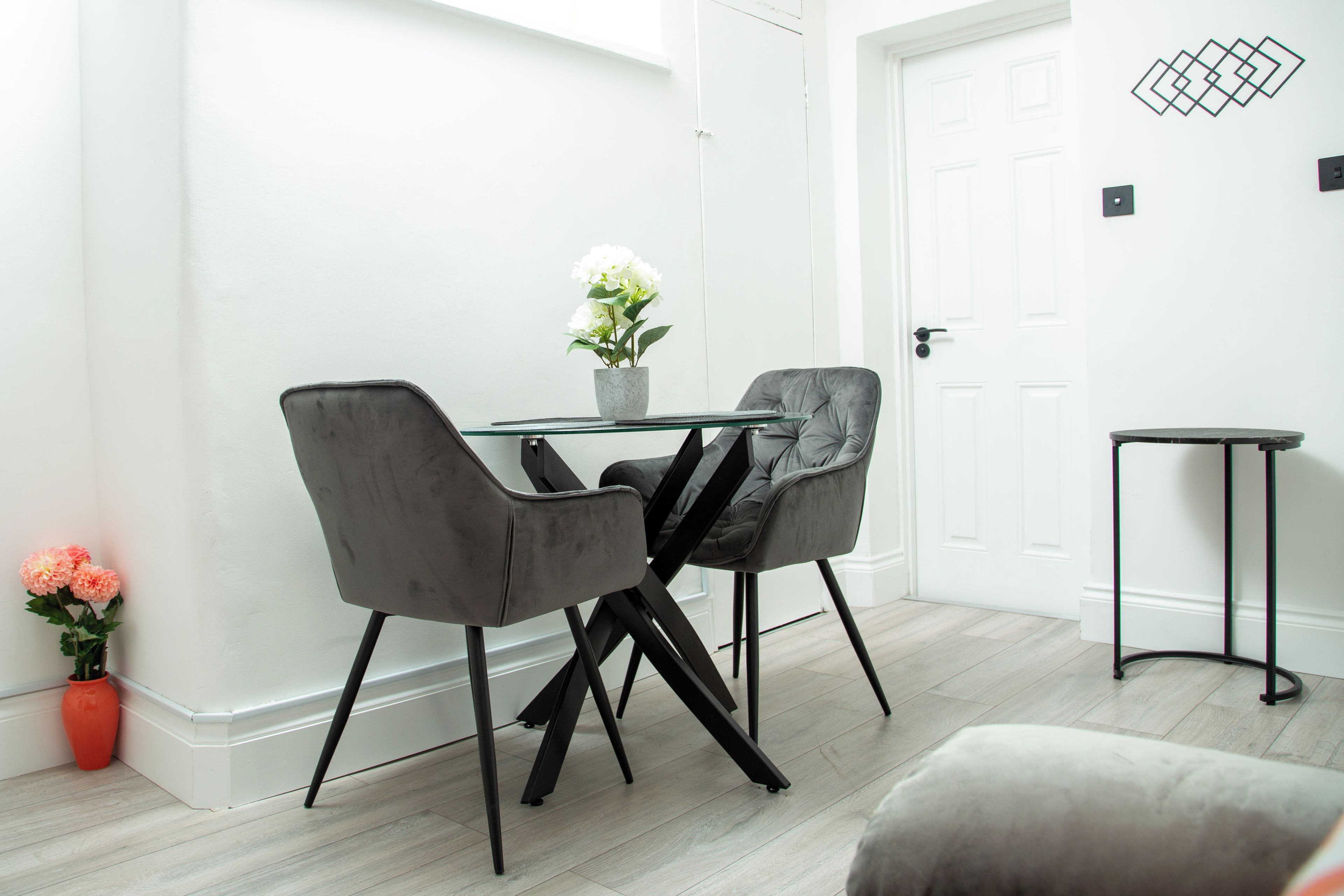 A modern dining area with a glass table, two grey upholstered chairs, and a potted plant.