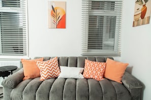 Ein graues Sofa mit gemusterten Kissen, ein kleiner runder Tisch und Kunstwerke an den Wänden.