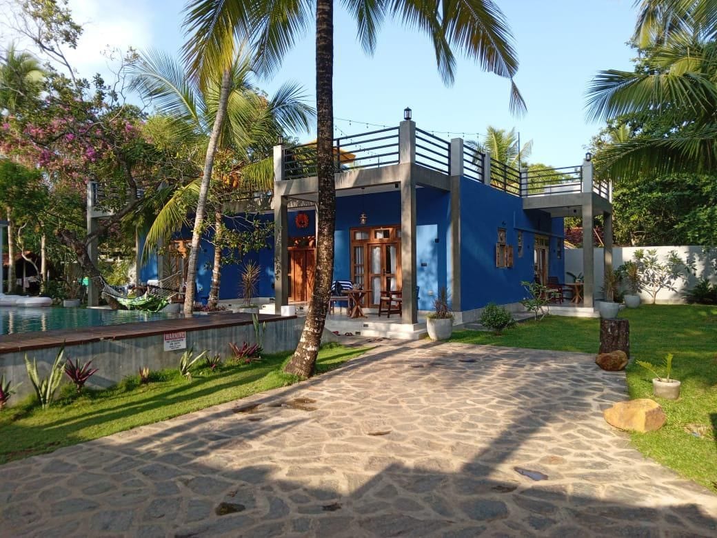 Un bâtiment à deux étages, à la façade bleue, avec un chemin de pierre menant à l’entrée et un espace piscine agrémenté d’un hamac.