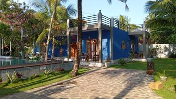 Un bâtiment à deux étages, à la façade bleue, avec un chemin de pierre menant à l’entrée et un espace piscine agrémenté d’un hamac.