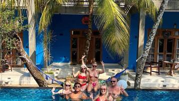 Un groupe de personnes dans une piscine, avec des palmiers et un bâtiment bleu en arrière-plan.