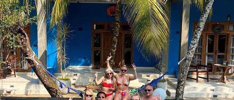 A group of people in a swimming pool with palm trees and a blue building in the background.