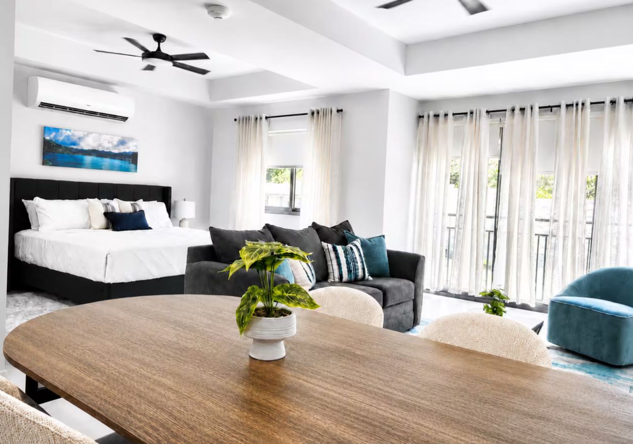 A modern hotel room with a bed, a sofa, a dining table, and a ceiling fan.