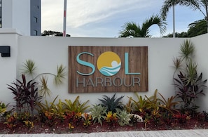 A sign for 'SOL HARBOUR' with a logo featuring a sun and wave, surrounded by various tropical plants. - Sol Harbour Apartment in Ocho Rios (Ocho Rios)