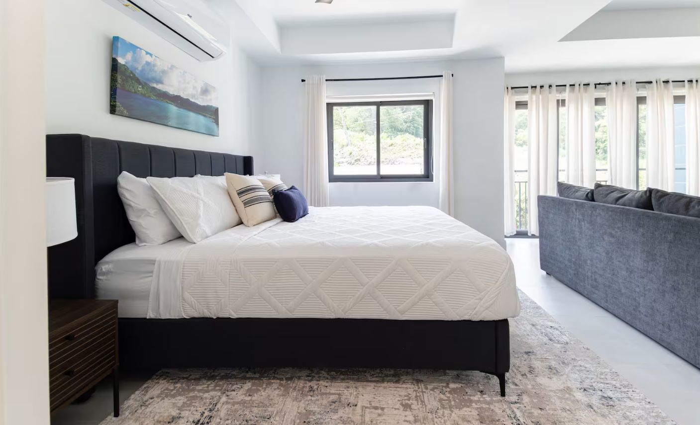 A modern bedroom with a large bed, a bedside table, a grey sofa, a window with curtains, and a painting on the wall.