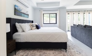 A modern bedroom with a large bed, a bedside table, a grey sofa, a window with curtains, and a painting on the wall. - Sol Harbour Apartment in Ocho Rios (Ocho Rios)