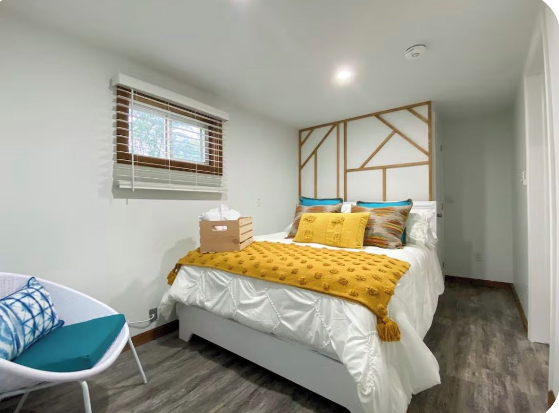 A bedroom with a bed, a chair, a window with blinds, and a wall with a geometric pattern.