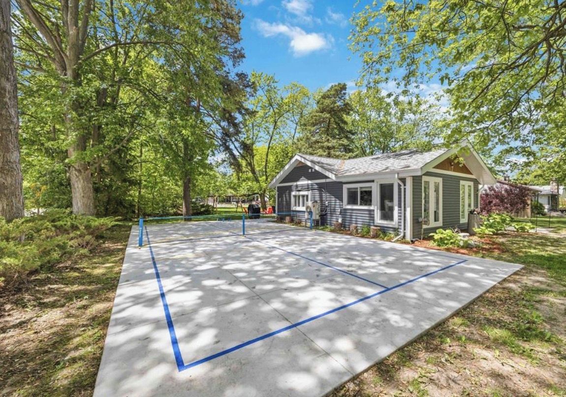 A residential property with a paved driveway, a small house with a gabled roof, and a yard with trees and shrubs.